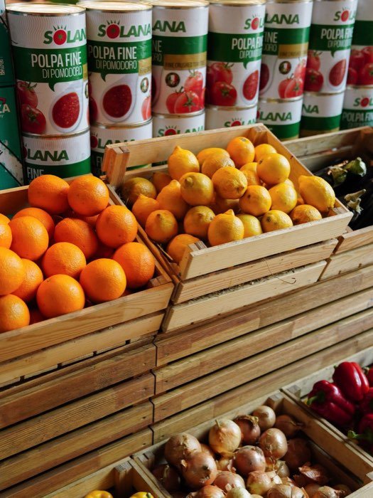 Wooden crates of oranges, lemons and onions stacked beside cases of tinned goods in a provisions store.